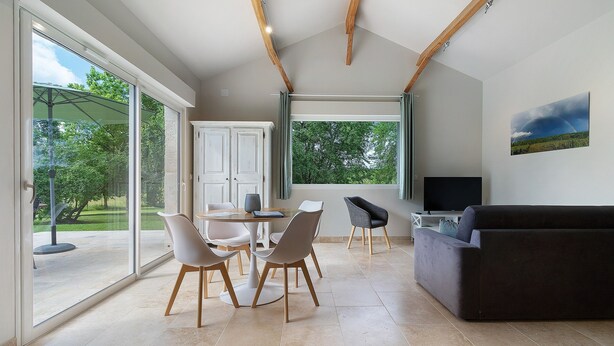 A spacious living area with a large sliding glass door leading to an outdoor patio, a dark-colored sofa, a dining table with chairs, and a flat-screen TV mounted on the wall. - Gîte L'albizia - Les Carassonnes with Shared Pool, Wi-Fi and Air Conditioning (Saint-Nexans)