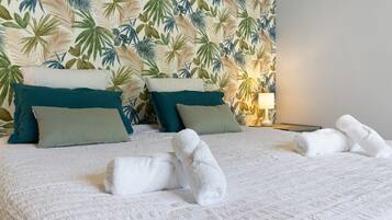 Un letto rifatto con lenzuola bianche e asciugamani arrotolati, una parete con motivo a foglia tropicale e una lampada da comodino.