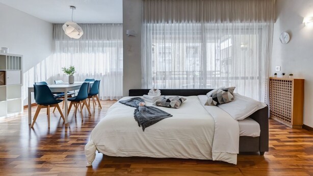 Moderni hotellihuone, jossa on suuri sänky, ruokailutila tuoleineen ja suuret ikkunat, joista avautuu näkymä kaupunkiin.