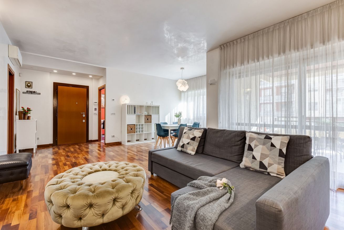 A modern living room with a sofa, ottoman, and a dining area in the background.