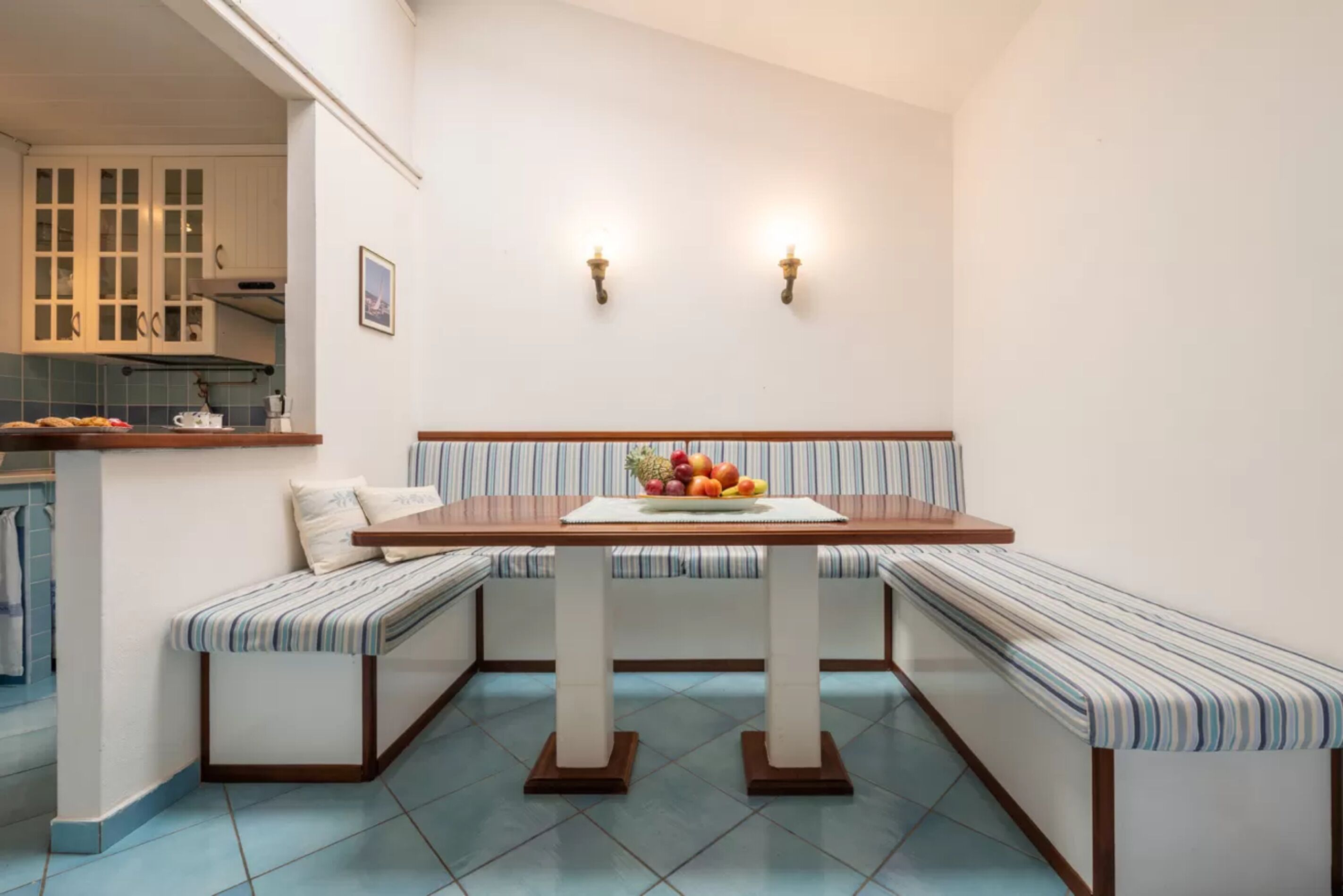 A corner kitchen with a bench seating area, a fruit bowl on the table, and a view of the kitchen in the background.