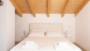 Une chambre avec un plafond en bois, un lit recouvert de draps blancs, deux tables de chevet et des lampes fixées au mur.