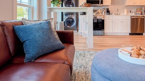 A modern living room with a leather sofa, a blue patterned pillow, and a round ottoman with snacks. - Charming 2BR•Walk to Harbor & Downtown•Close2Salem (Gloucester)