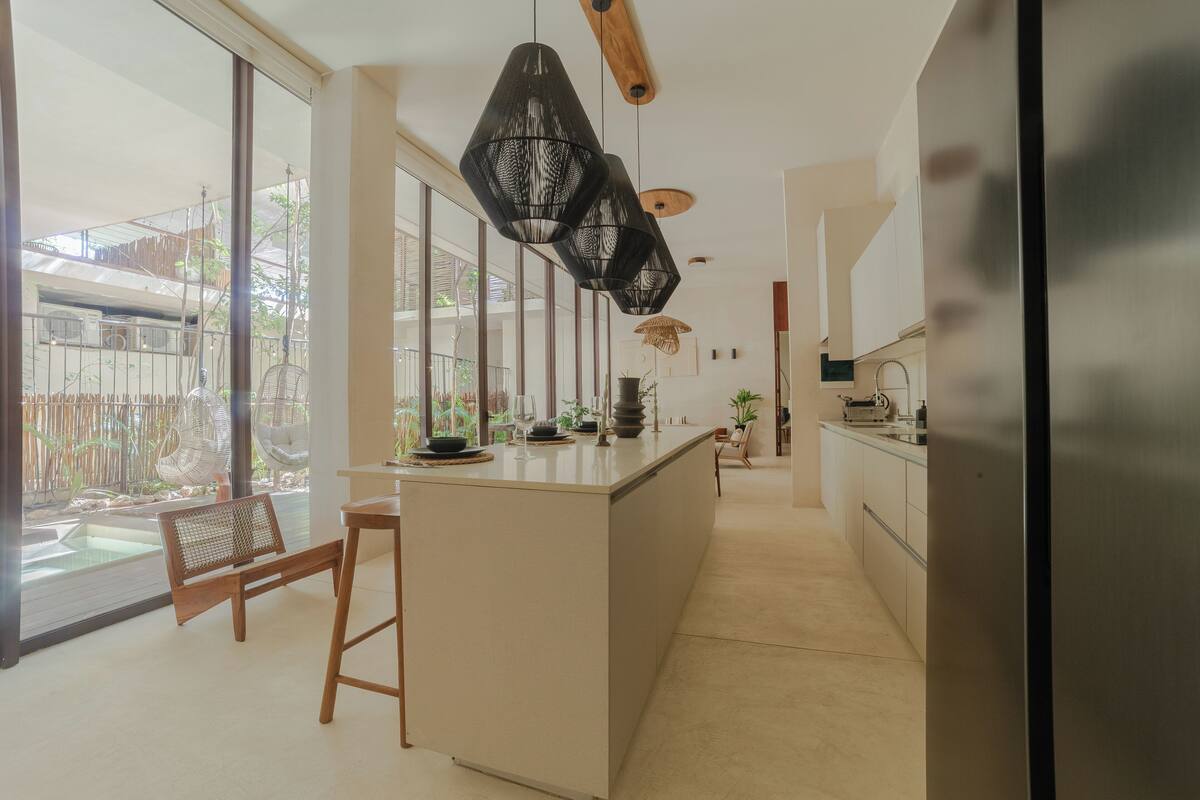 A modern kitchen with large windows, a central island, and hanging pendant lights.