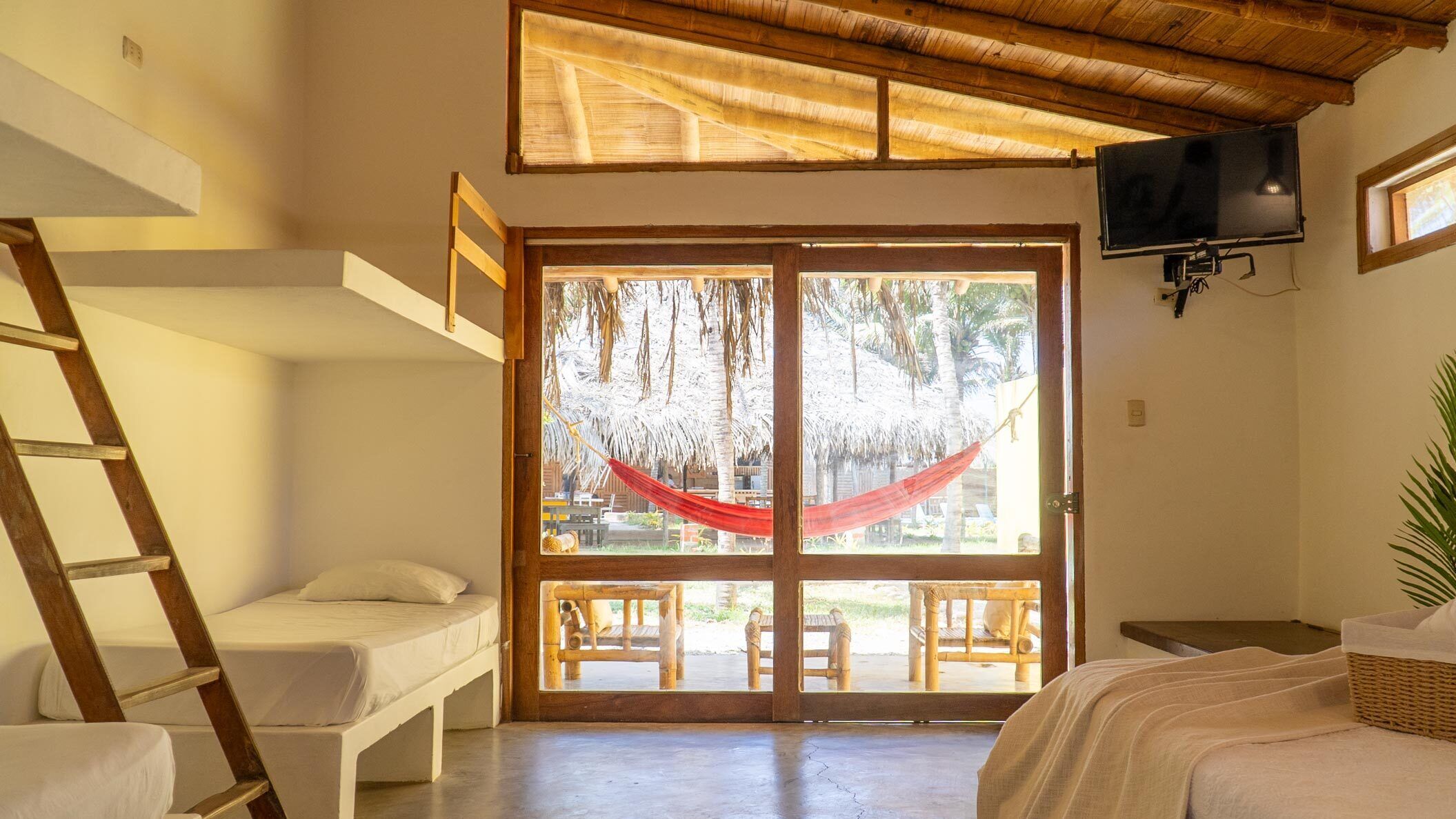 A room with a bed, a wooden ladder, a TV mounted on the wall, and a large sliding glass door leading to an outdoor area with a hammock and wooden furniture.