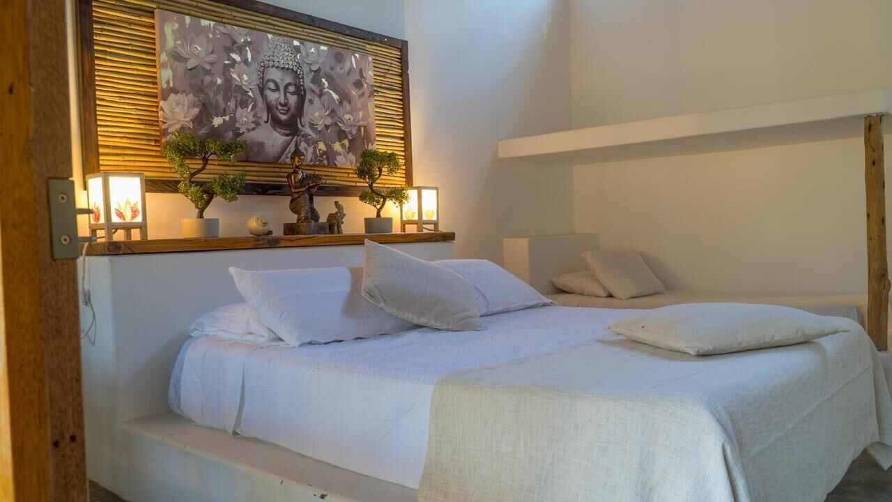 A bedroom with a bed, pillows, a nightstand, a wall-mounted shelf with decorative items, and a framed artwork above the bed.