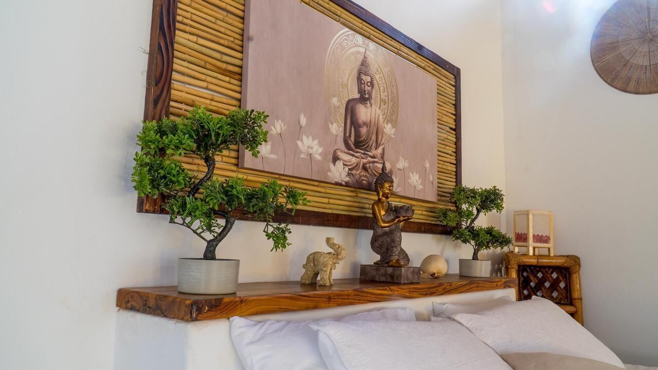 A bedroom with a bed, a wooden shelf with decorative items, a framed Buddha painting, and a small Buddha statue.
