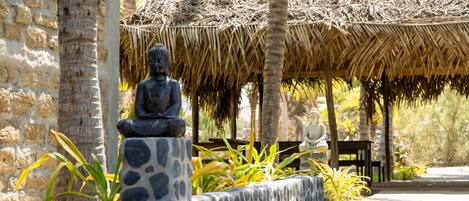 A stone Buddha statue in a garden with a thatched roof structure in the background.
