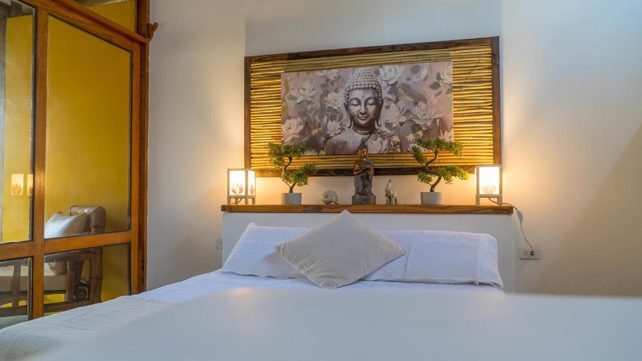 Una cama bien hecha con sábanas blancas, una estatua de Buda y dos plantas en macetas sobre una mesita de noche.