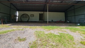 Un almacén amplio y abierto con techo metálico y paredes corrugadas. En su interior, hay puertas de metal, un objeto circular grande y varios objetos dispersos.