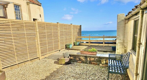 A small outdoor patio area with a wooden fence, a stone wall, and a bench. There are potted plants and a small table. The background features a view of the sea and a building with a tiled roof. - Little Net House | Pittenweem (Pittenweem)