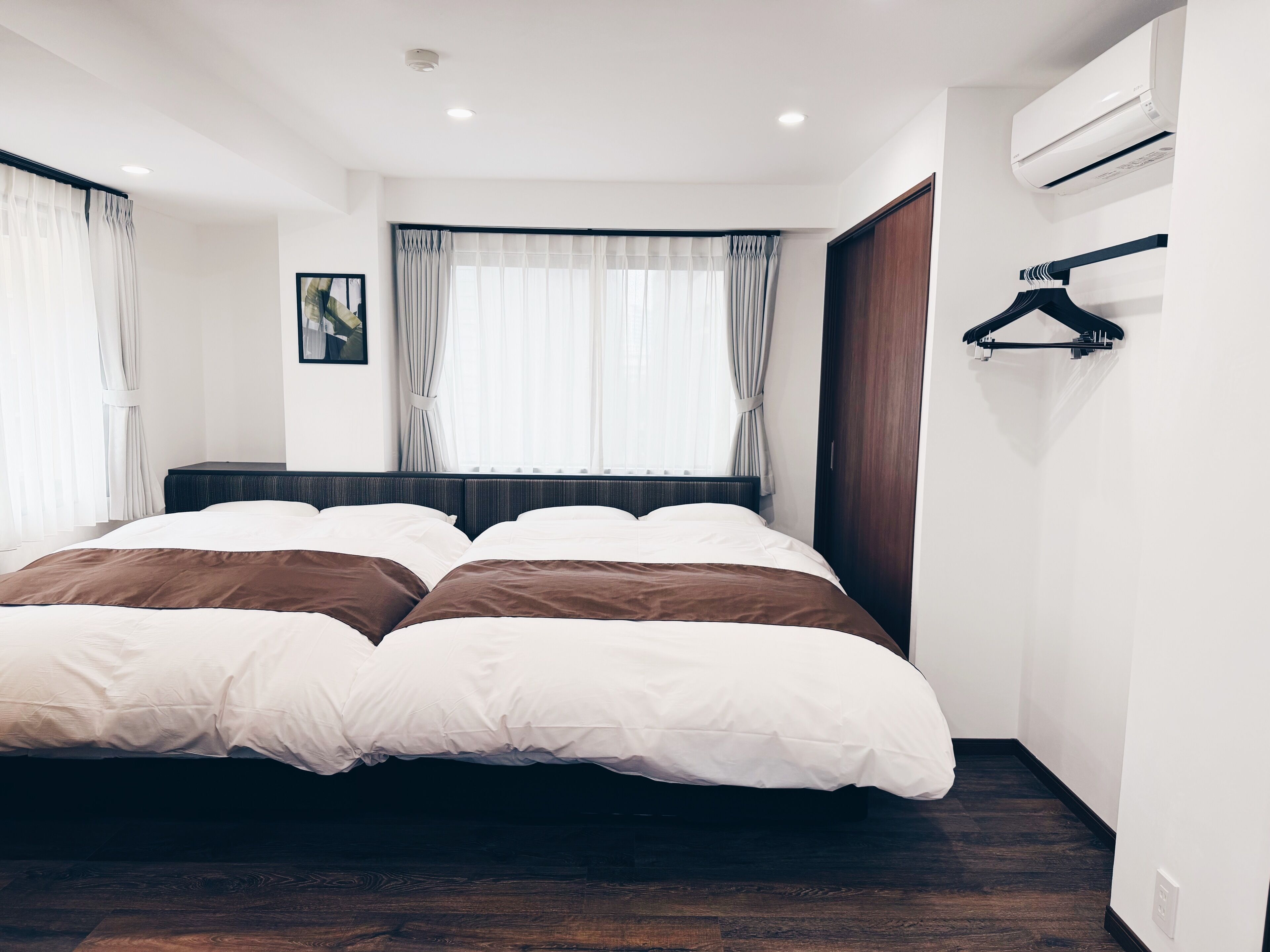 A modern hotel room with a large bed, a wooden floor, a window with curtains, and an air conditioning unit.