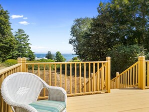 A wooden deck with a wicker chair, overlooking a coastal view with trees and a clear sky.