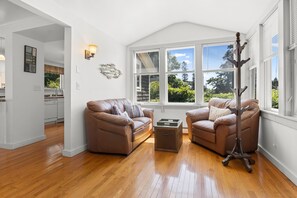 A spacious living room with a high ceiling, a large window with blinds, a brown leather sofa, a matching armchair, and a coat rack.