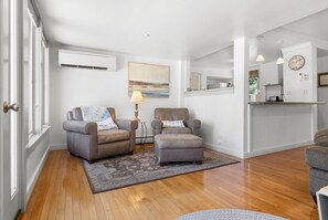 A living room with two armchairs, a rug, a painting, and a kitchen area with a clock and a window.