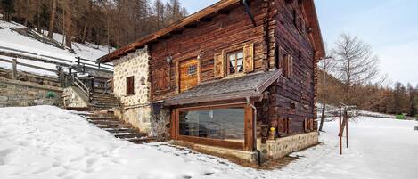 Une cabane en rondins de bois, avec des fondations en pierre, une grande fenêtre et un escalier menant à l’entrée.