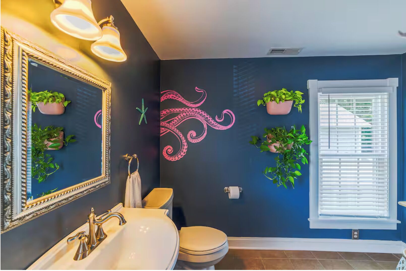 A bathroom with a dark blue wall, a large mirror, a sink, a toilet, and potted plants.