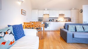 Ein modernes Wohnzimmer mit einer Couch, einem Esstisch mit Stühlen, einer Küchenzeile mit weißen Schränken und einer Uhr an der Wand.