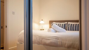 Ein ordentlich bezogenes Bett mit weißer Decke und gestreifter Bettwäsche, eine Nachttischlampe und eine Steckdose an der Wand.