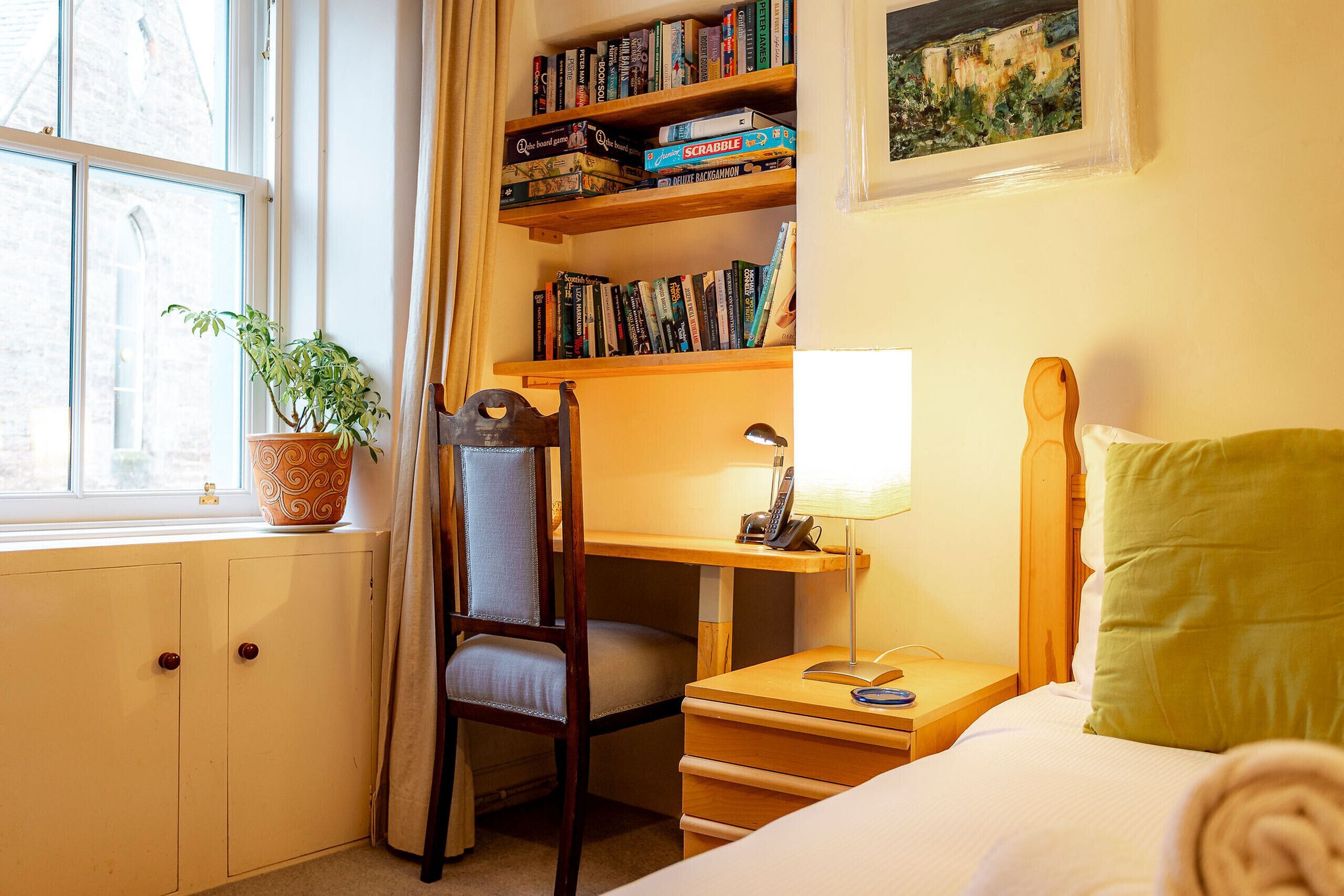 A hotel room with a bed, a desk with a lamp, a chair, a bookshelf with books, a potted plant, and a window with curtains.