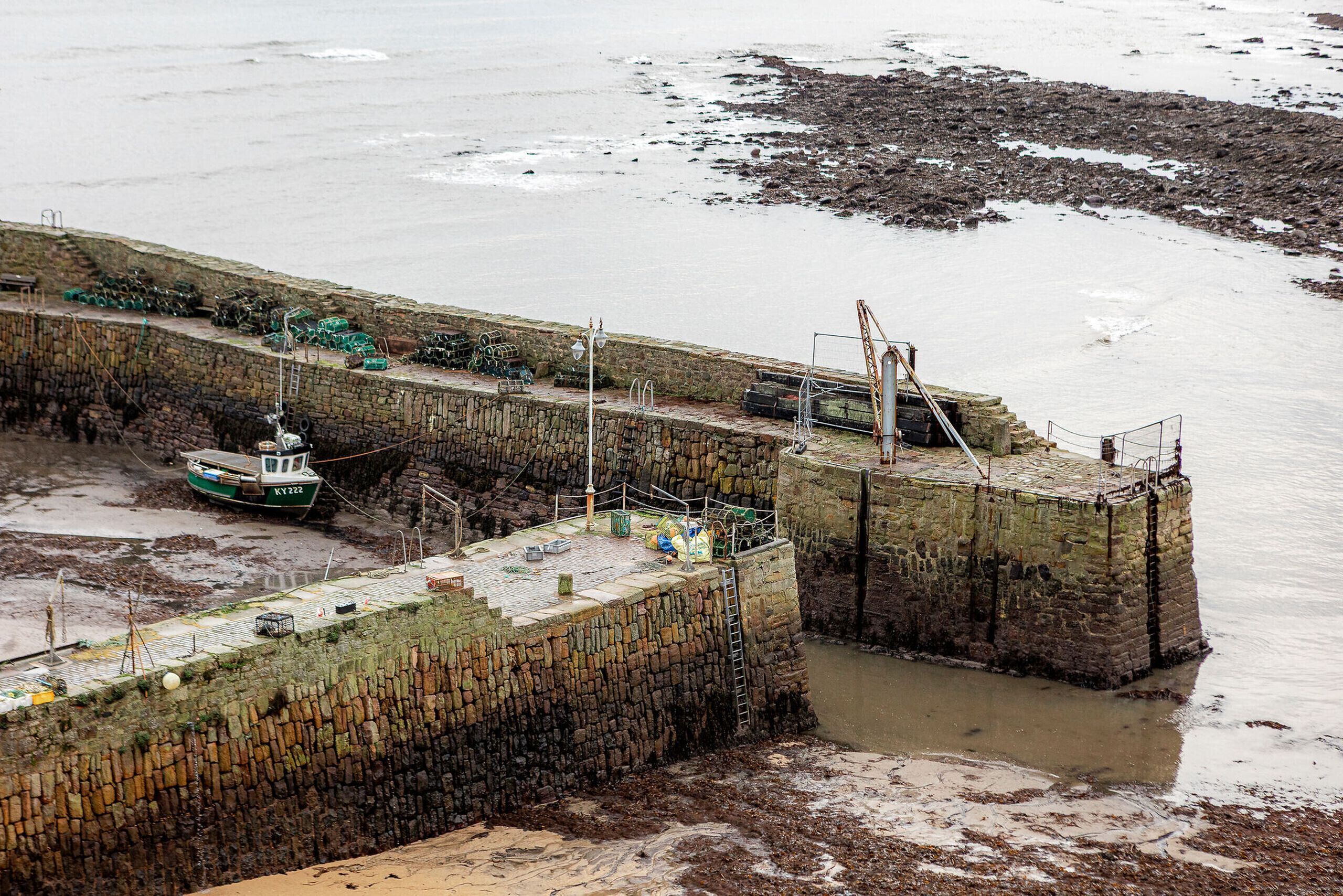 A coastal pier with boats docked, a stone wall, and a rocky shoreline.