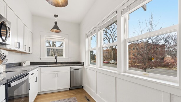 A modern kitchen with white cabinetry, a stainless steel sink, and a dishwasher. There are large windows providing a view of the street and buildings outside. - Mathews House 4 King Beds Near CSU & Old Town (Fort Collins)