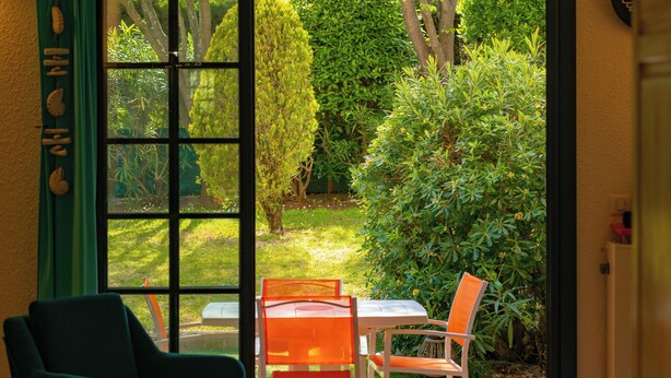 An open door leading to a patio with a table and two chairs, a green chair inside, and a view of a garden with various plants and a tree. - Very nice app 150m from the beach 50m from the pool (Saint-Raphaël)