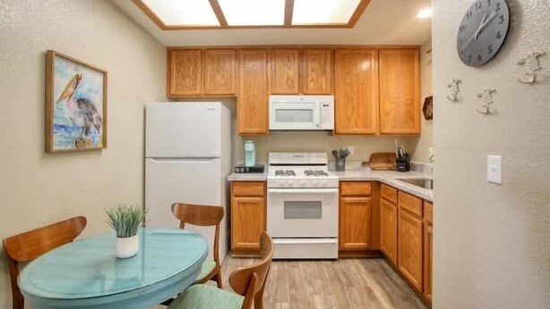 Cocina compacta con muebles de madera, un refrigerador blanco, un microondas y una estufa. Hay una mesa de comedor con sillas y una pequeña planta en maceta.