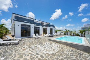 A modern house with a swimming pool, stone patio, and white lounge chairs.