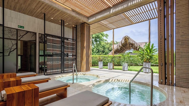 Een wellnessruimte met houten ligbedden, een jacuzzi en uitzicht op een gebouw met rieten dak.