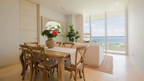 Een eetruimte met een houten tafel en stoelen, een vaas met bloemen en uitzicht op zee door grote ramen.