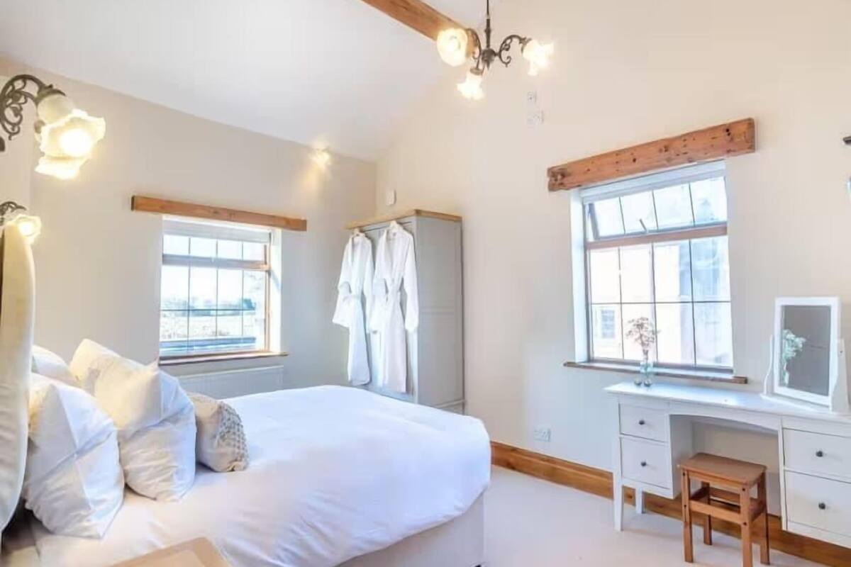 A bedroom with a bed, a desk, and a chair. There are windows with curtains, a wardrobe, and a chandelier.
