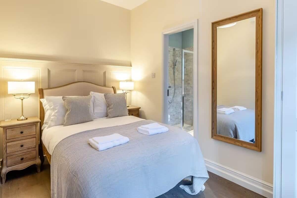 A neatly arranged bedroom with a bed, bedside lamps, a mirror, and a shower area.