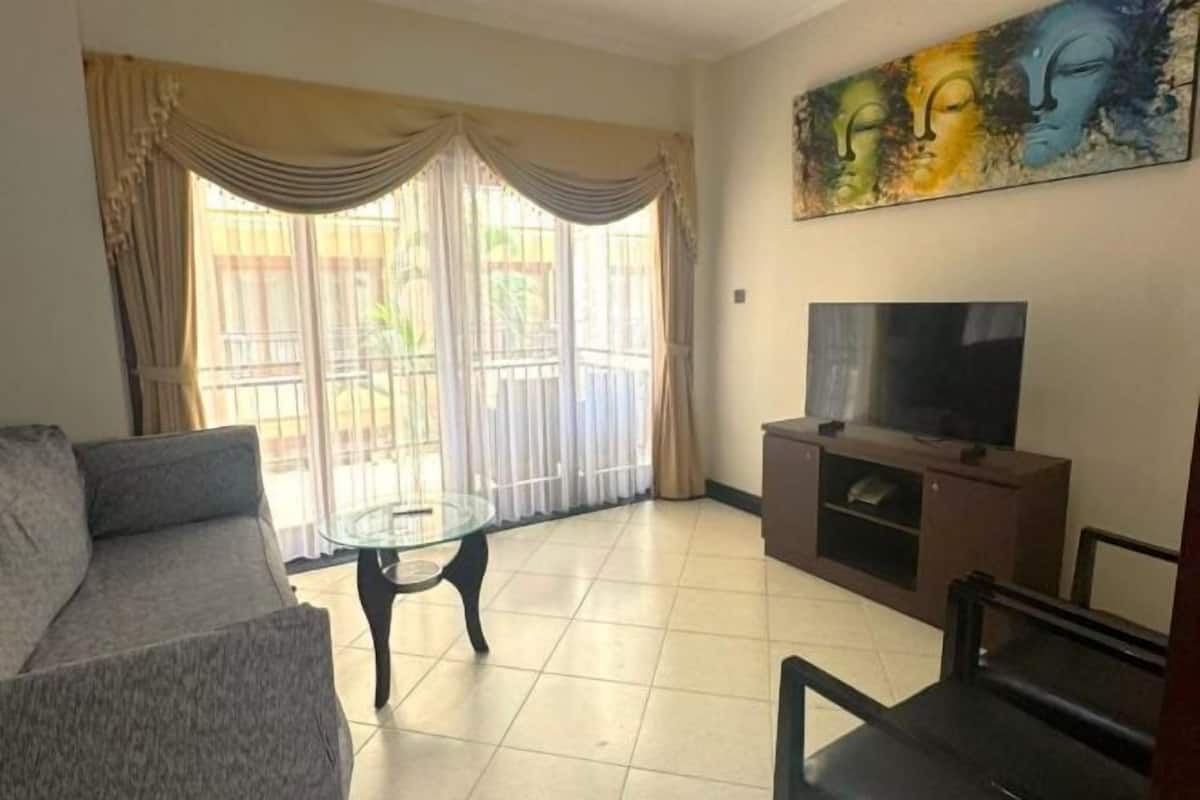 A living room with a sofa, a glass coffee table, a TV on a wooden stand, and a painting on the wall.