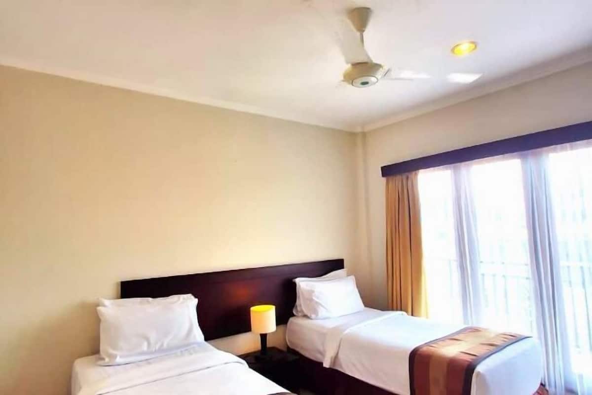 A hotel room with two beds, a ceiling fan, a window with curtains, and a nightstand with a lamp.