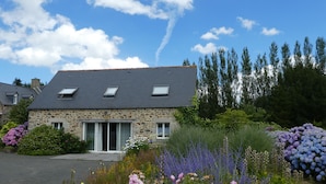 Une maison en pierre avec un toit en pente, des fenêtres de toit et un jardin où poussent différentes sortes de fleurs.
