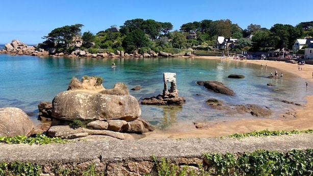 Une plage côtière avec une eau claire, de gros rochers et quelques personnes.
