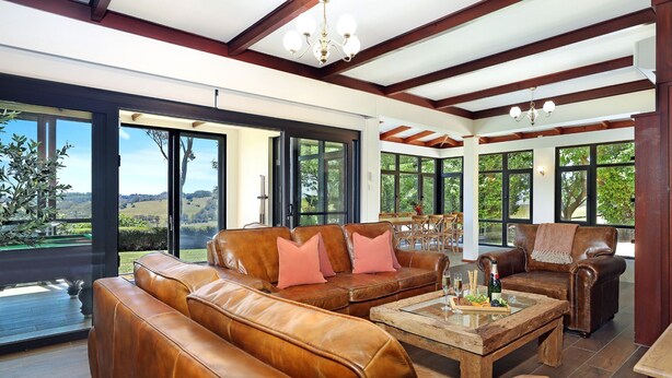A spacious living room with leather sofas, a wooden coffee table, and large windows offering a view of the countryside. - Goolara – luxurious two-storey escape with ensuite bedrooms, sauna and countryside views (Burrawang)