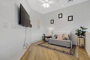 Ein modernes Wohnzimmer mit einem wandmontierten Flachbildfernseher, einem grauen Sofa mit gemusterten Kissen, einem Holzboden und einem Deckenventilator.