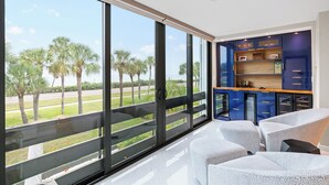 Une chambre moderne dotée d’une grande porte-fenêtre coulissante donnant sur un balcon, d’un coin salon avec des fauteuils à motifs et d’une cuisine intégrée avec des meubles bleus.