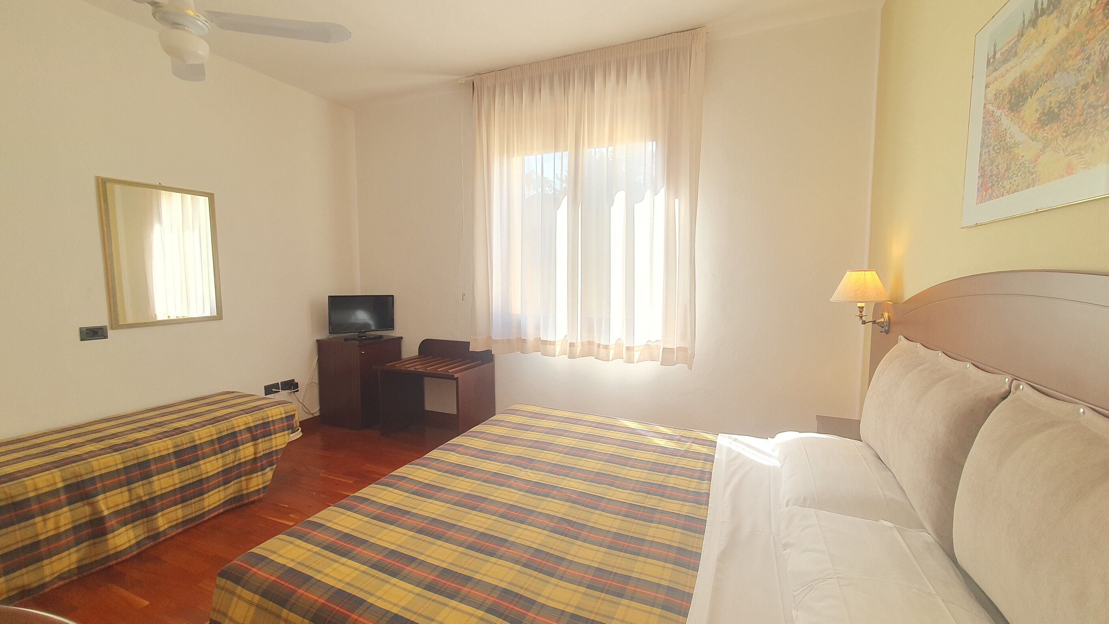 A hotel room with a bed, a desk with a television, a chair, a bench, and a window with curtains.