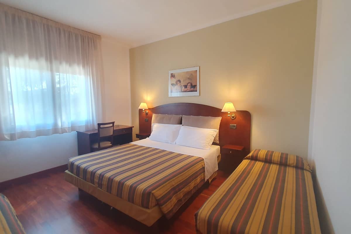 A hotel room with a double bed, two bedside tables, a desk, a chair, and a window with curtains.
