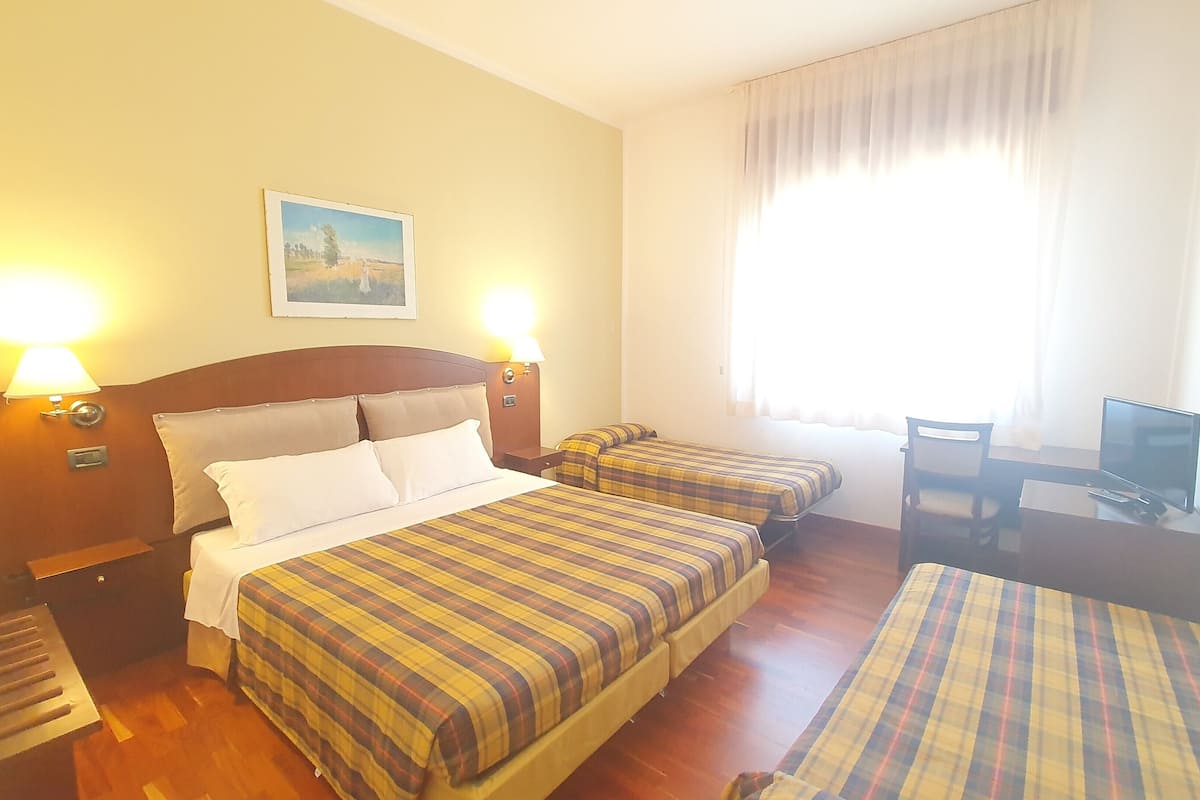 A hotel room with a bed, two bedside tables, a desk with a chair, a television, and a window with curtains.