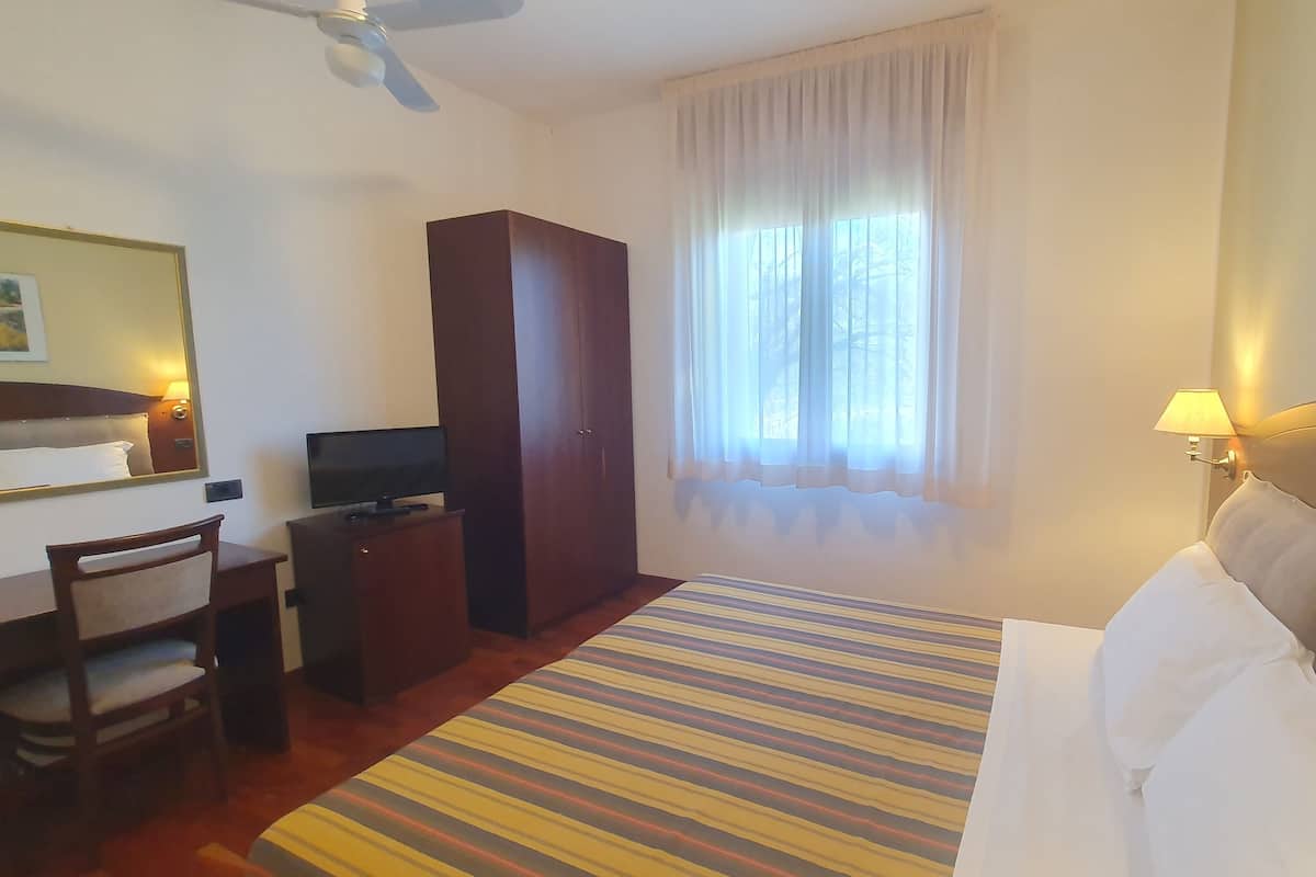 A hotel room with a bed, a desk with a television, a wardrobe, a window with curtains, and a ceiling fan.