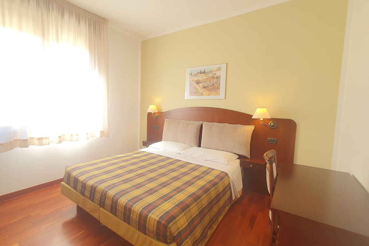 A hotel room with a bed, wooden headboard, two bedside lamps, a nightstand, a window with curtains, and a painting on the wall.