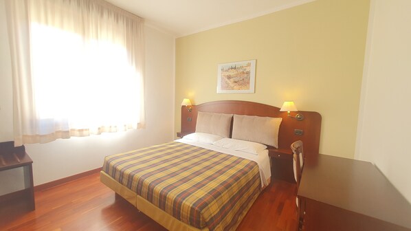 Habitación de hotel con cama, cabecera de madera, dos lámparas de noche, una mesita de noche, una ventana con cortinas y un cuadro en la pared.