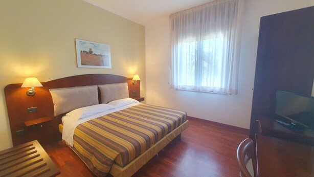 A hotel room with a bed, two bedside lamps, a painting on the wall, a window with curtains, and a television on a stand. - Agriturismo Gli Ulivi (Predappio)