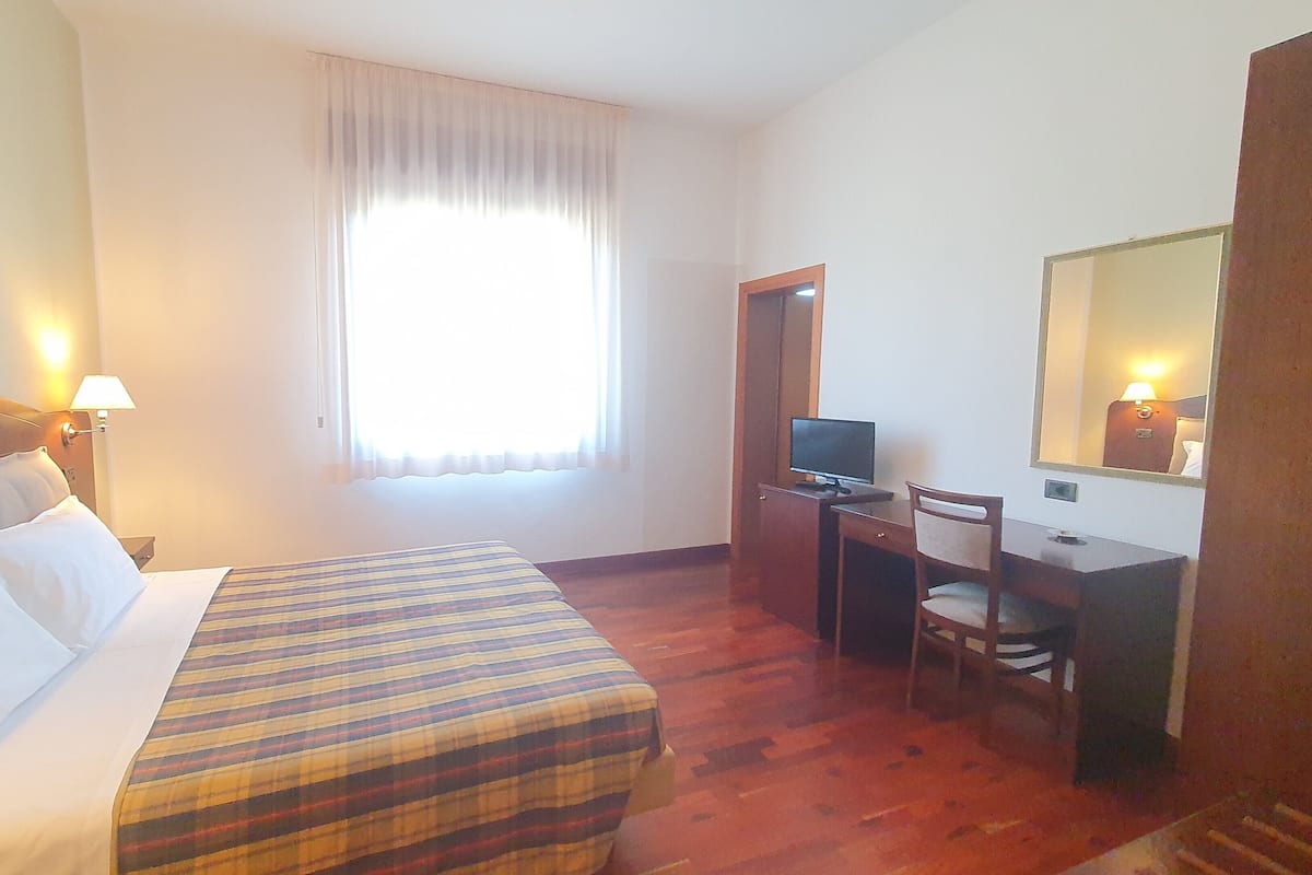 A hotel room with a bed, a desk with a computer, a chair, and a large window with curtains.