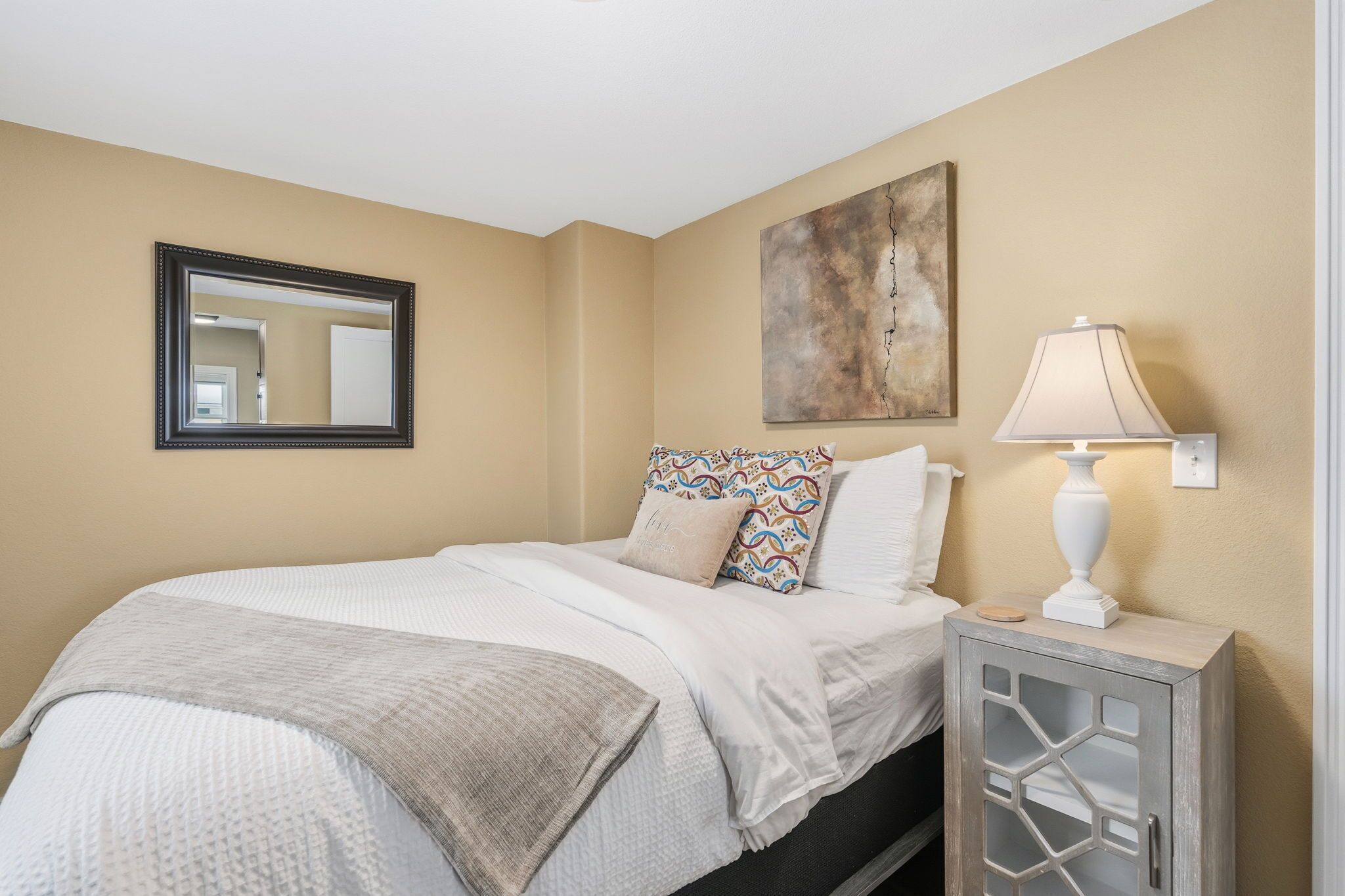 A neatly made bed with patterned pillows, a bedside lamp, and a wall-mounted mirror.