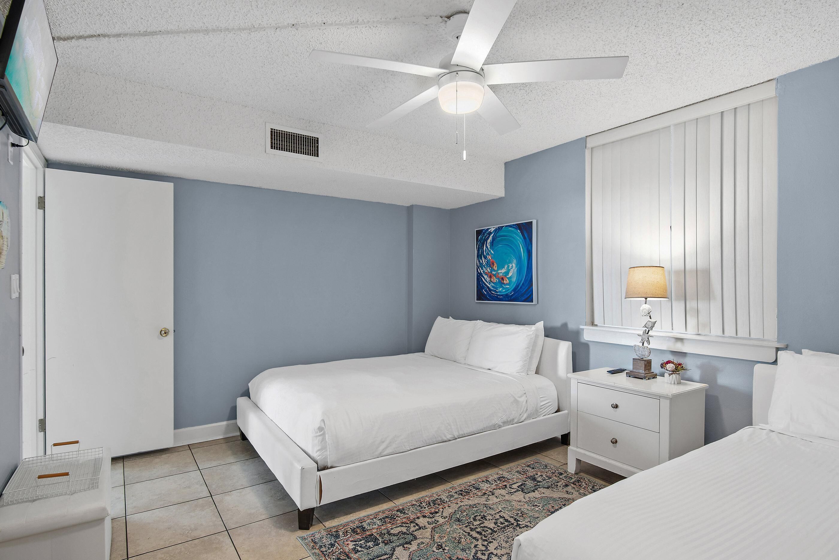 Ein Schlafzimmer mit Bett, Nachttisch, Lampe, einem Fenster mit Jalousien und Deckenventilator.